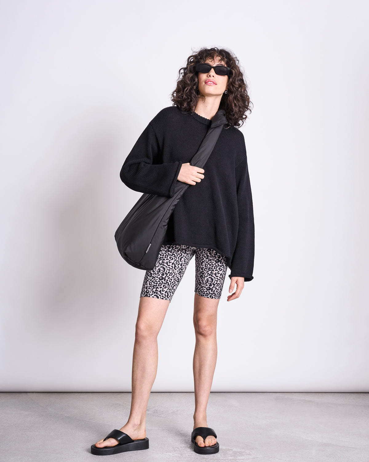 A woman with curly hair wears jan-n-junes BIKER SHORTS LEO GOTS, black sunglasses, a loose black sweater, slide sandals, and a shoulder bag, standing against a light background.