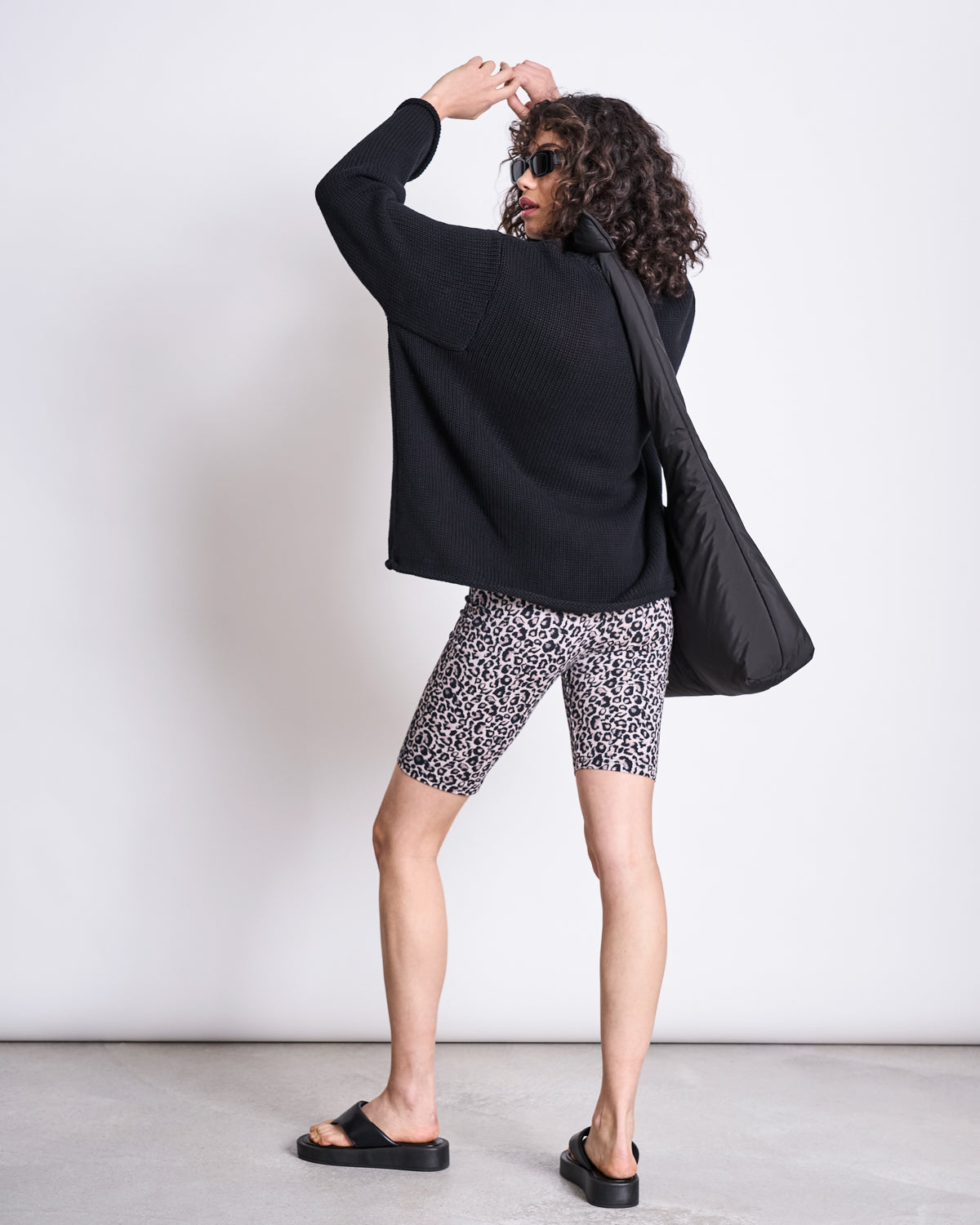 A person with curly hair in sunglasses, a black oversized sweater, leopard print bike shorts, and black sandals carries the jan-n-june BAG BIWA PUFFY REGULAR BLACK—water-repellent and made from recycled plastic—against a plain background.