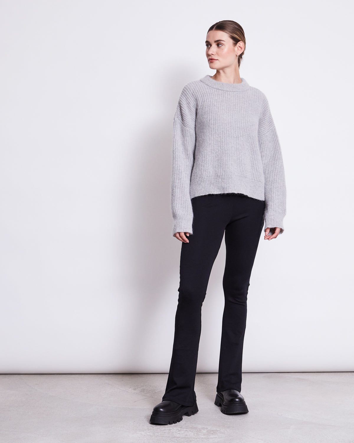 A person stands against a plain white wall, wearing jan-n-june FLARED LEGGINGS BLACK, a loose light gray TENCEL Lyocell sweater, and black platform shoes, looking slightly to the side with hands relaxed by their sides.