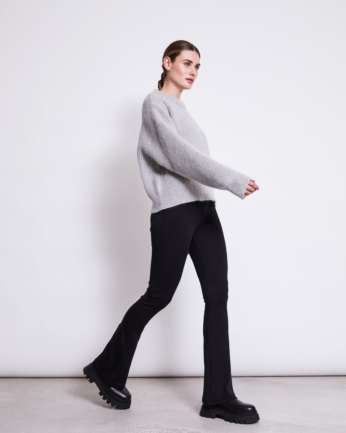 A woman with her hair pulled back and a neutral expression walks sideways in front of a white wall, wearing jan-n-junes FLARED LEGGINGS BLACK, a loose grey organic cotton sweater, and chunky black boots.