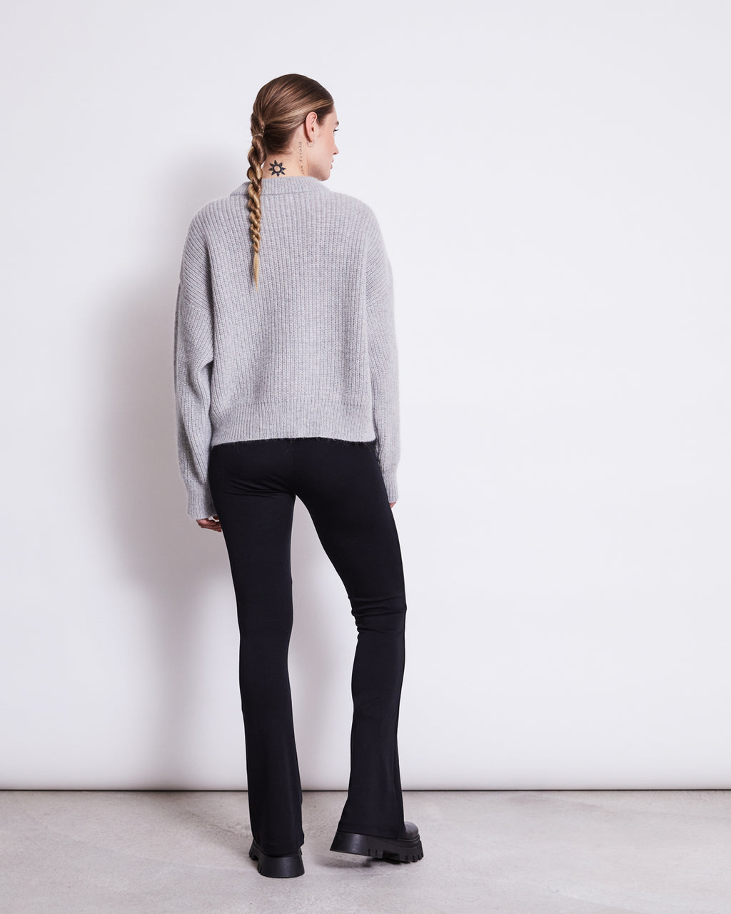 A person with a long braid faces away, wearing a light gray sweater and jan-n-june FLARED LEGGINGS BLACK, made from soft organic cotton, paired with chunky black shoes against a plain white wall.