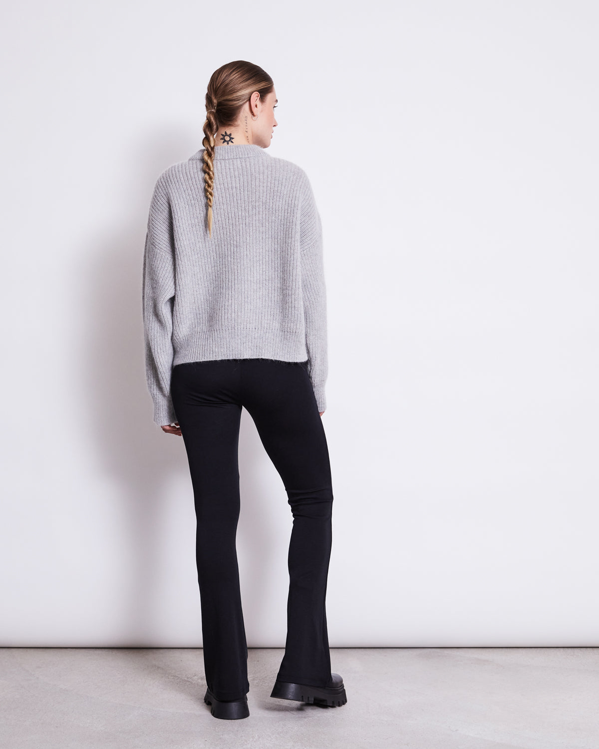 A person with a long braid faces away, wearing a light gray sweater and jan-n-june FLARED LEGGINGS BLACK, made from soft organic cotton, paired with chunky black shoes against a plain white wall.
