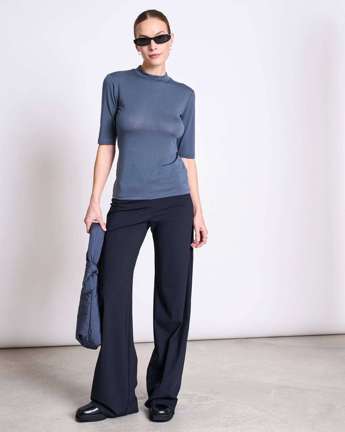 A woman stands against a plain background, wearing sunglasses, wide-leg black pants, black shoes, and the jan-n-june LYOCELL HIGH-NECK T-SHIRT TARMA TURBULENCE. She holds a blue-gray quilted bag at her side.