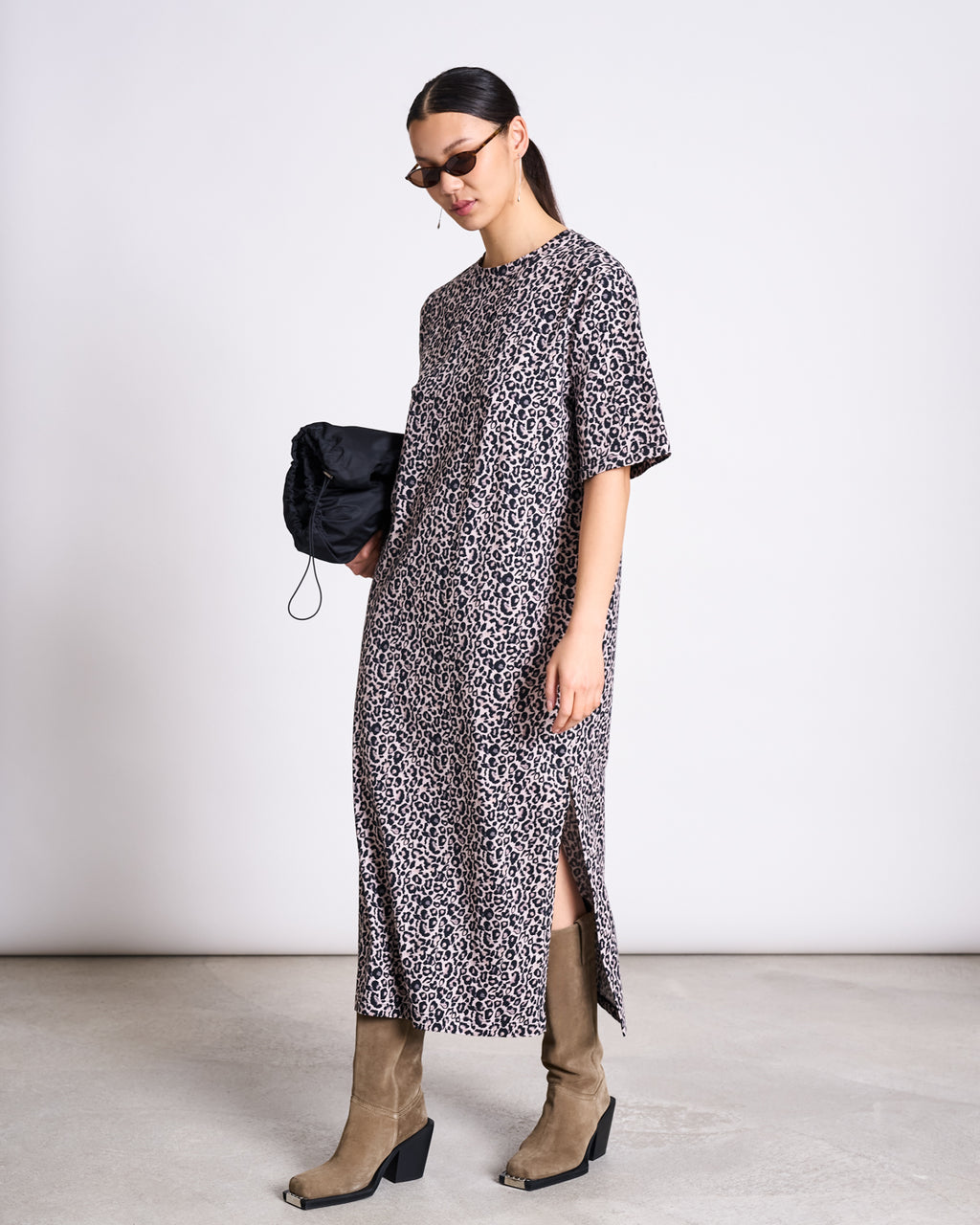 A woman wearing the jan-n-june SHIRT DRESS CROVIE LEO PRINT GOTS with tan knee-high boots and sunglasses stands against a plain background, holding a black bag at her side.