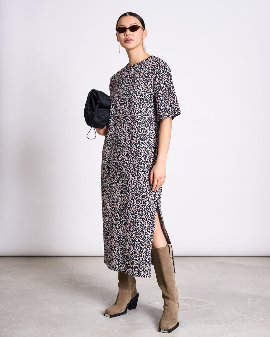 A woman wearing sunglasses and a jan-n-june SHIRT DRESS CROVIE LEO PRINT GOTS with tan knee-high boots holds a small black bag, standing against a plain white wall.
