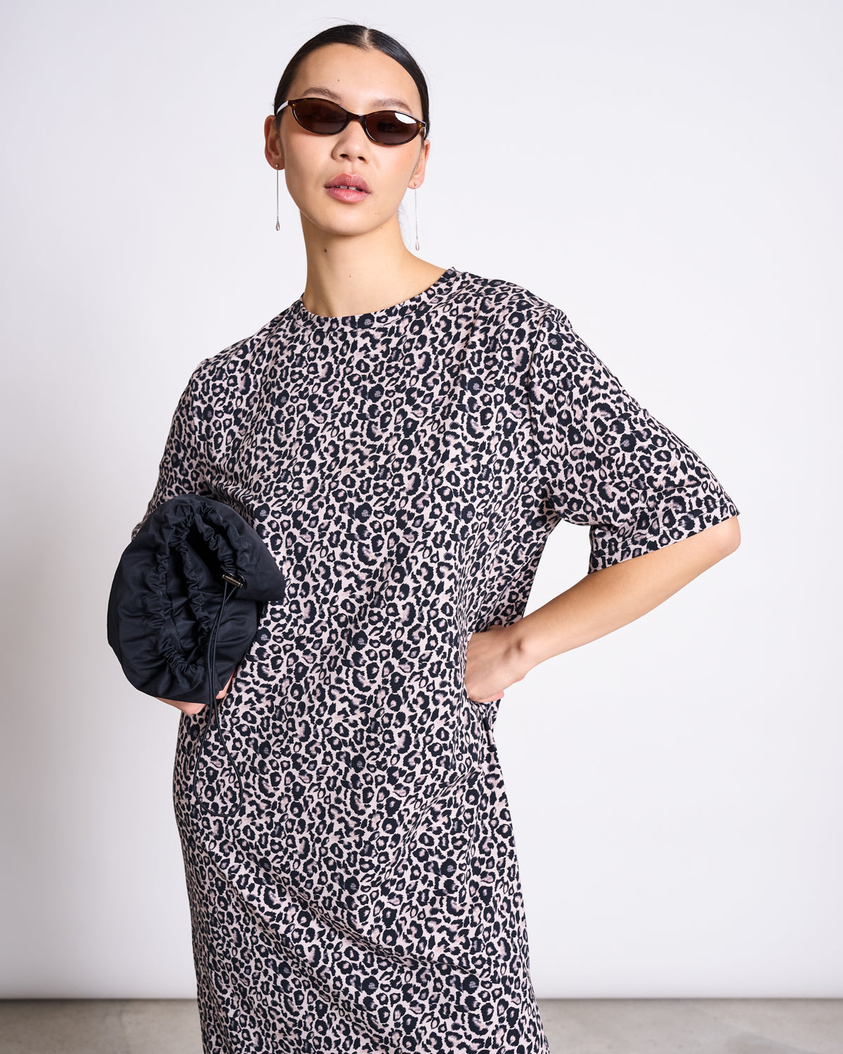 A person wears the jan-n-june SHIRT DRESS CROVIE LEO PRINT GOTS—a loose, short-sleeved, leopard print midi dress—along with brown sunglasses and drop earrings, holding a black ruched clutch against a plain background.