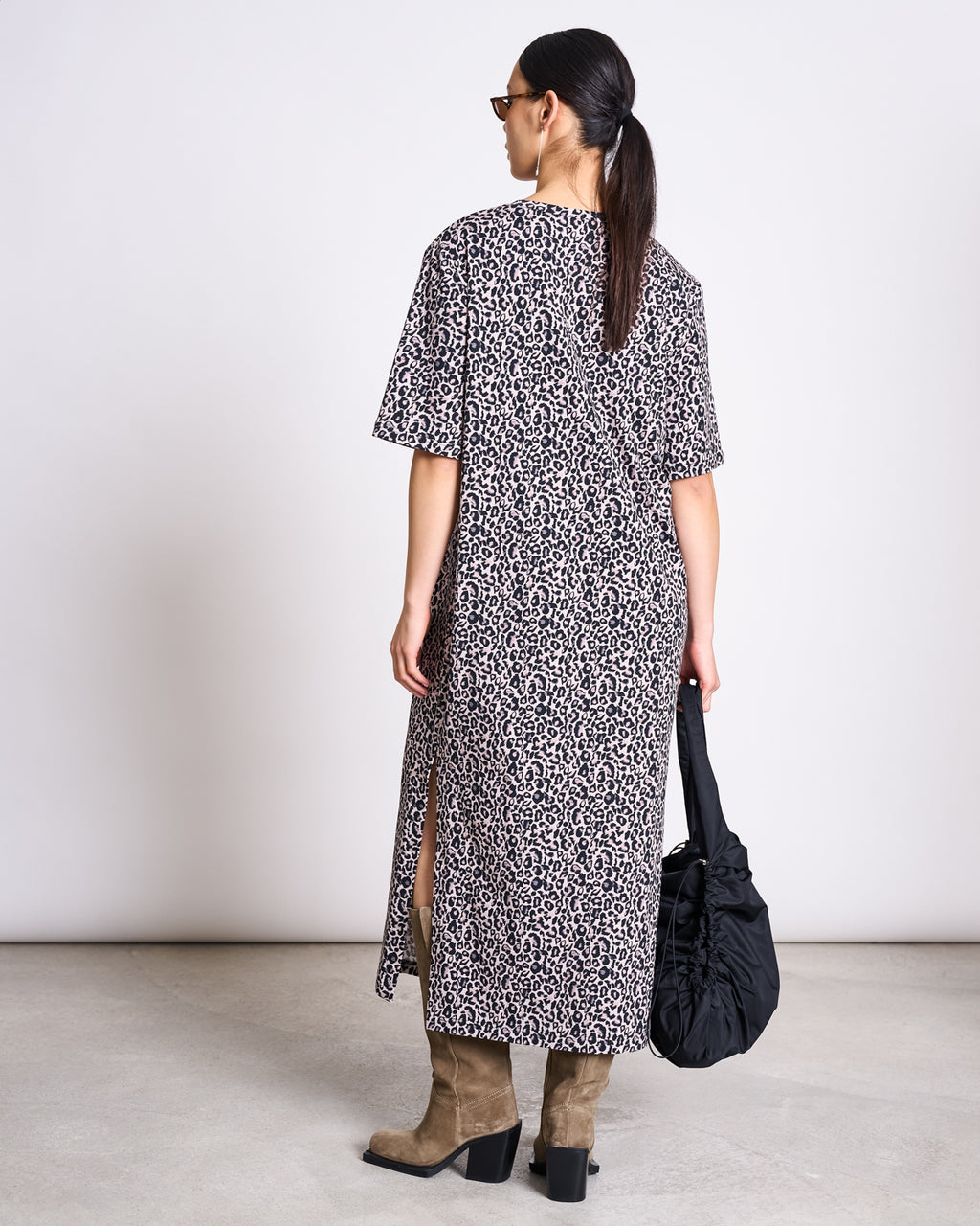 A woman with a ponytail and sunglasses wears the jan-n-june SHIRT DRESS CROVIE LEO PRINT GOTS, paired with taupe suede boots, as she stands facing away holding a black ruched bag in her left hand.