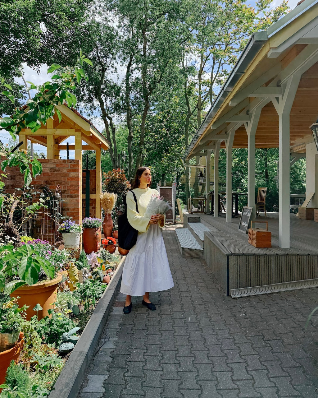 A woman wears the jan-n-june JUMPER SONA BUTTERMILK GOTS, standing on a garden path with flowers, potted plants and a wooden gazebo to her left, and a covered porch to her right on a sunny day.
