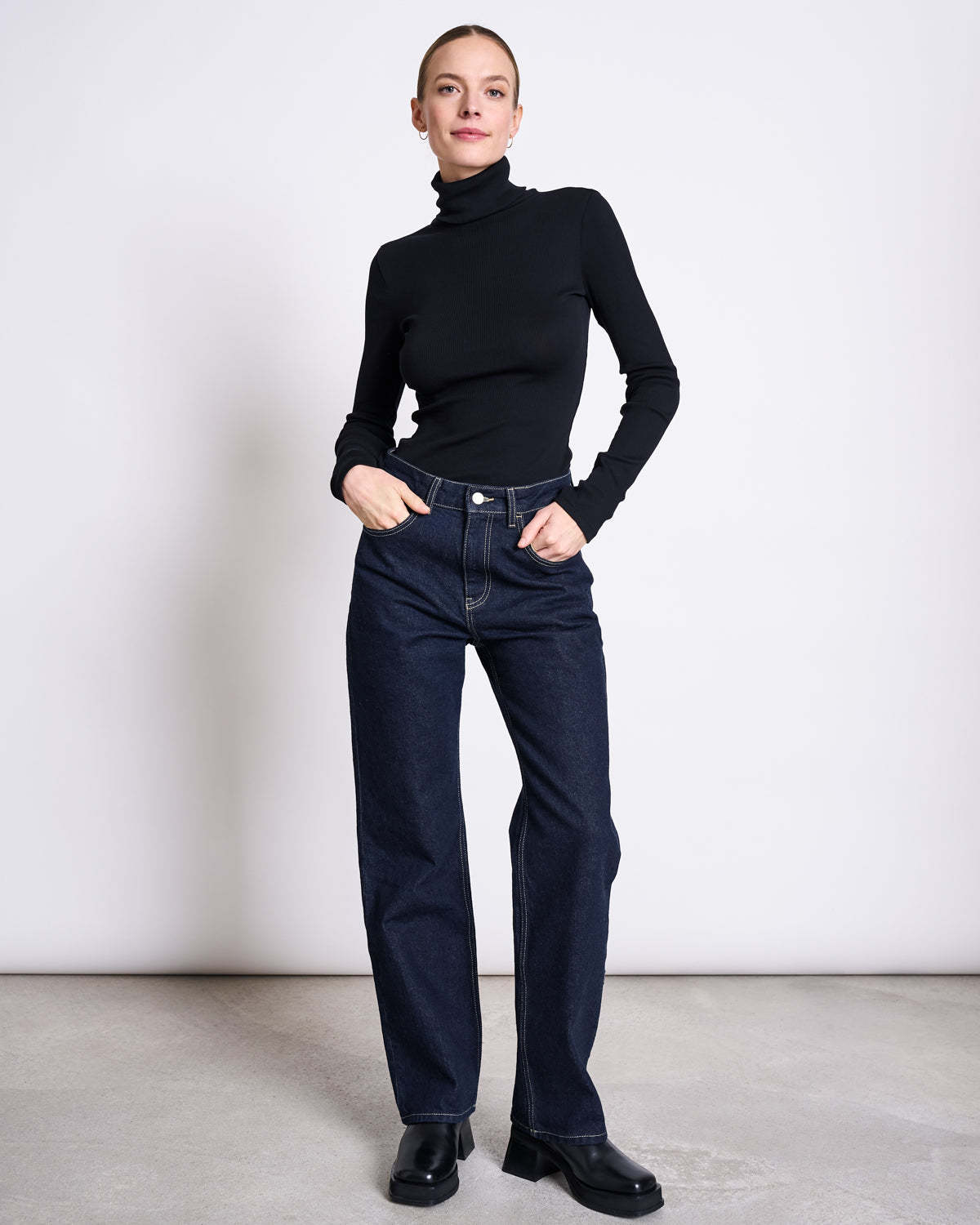 A woman stands against a white background, smiling slightly with her hair pulled back. She wears a fitted black turtleneck, jan-n-junes STRAIGHT LEG HIGHWAIST JEANS VILCA DARK BLUE DENIM GOTS, and black shoes.