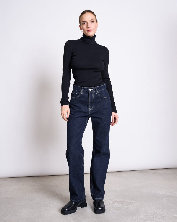 Indoors against a plain wall, a person wears the jan-n-june MINIRIB TURTLENECK CHUVA BLACK GOTS with dark blue high-waisted jeans and black shoes. Their hair is pulled back, with a neutral expression and arms relaxed by their sides.