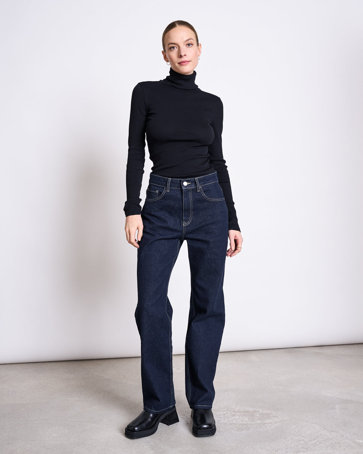 A woman stands against a plain light wall, wearing a fitted black turtleneck, jan-n-june STRAIGHT LEG HIGHWAIST JEANS VILCA DARK BLUE DENIM GOTS, and black shoes. Her hair is tied back as she looks at the camera with a neutral expression.