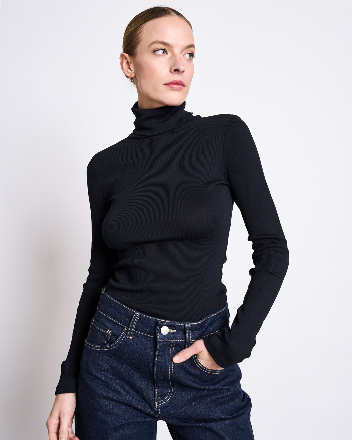 A woman in a fitted black turtleneck and jan-n-june STRAIGHT LEG HIGHWAIST JEANS VILCA DARK BLUE DENIM GOTS stands against a white background, gazing to the side with one hand in her pocket.