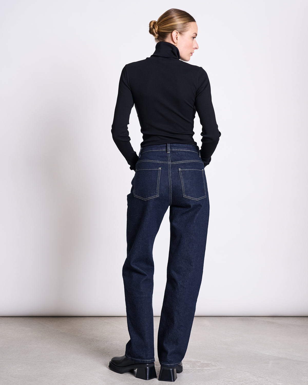 A person stands facing away against a white background, wearing jan-n-june STRAIGHT LEG HIGHWAIST JEANS VILCA DARK BLUE DENIM GOTS, a black turtleneck, black shoes, and has their hair tied back. White stitching on the jeans is visible.