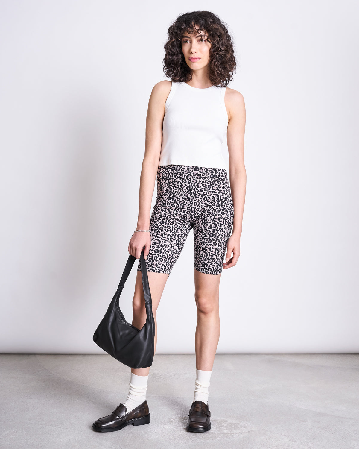 A woman with curly hair wears jan-n-junes BIKER SHORTS LEO GOTS, a white sleeveless top, white socks, black loafers, and carries a black handbag while standing against a plain light background.