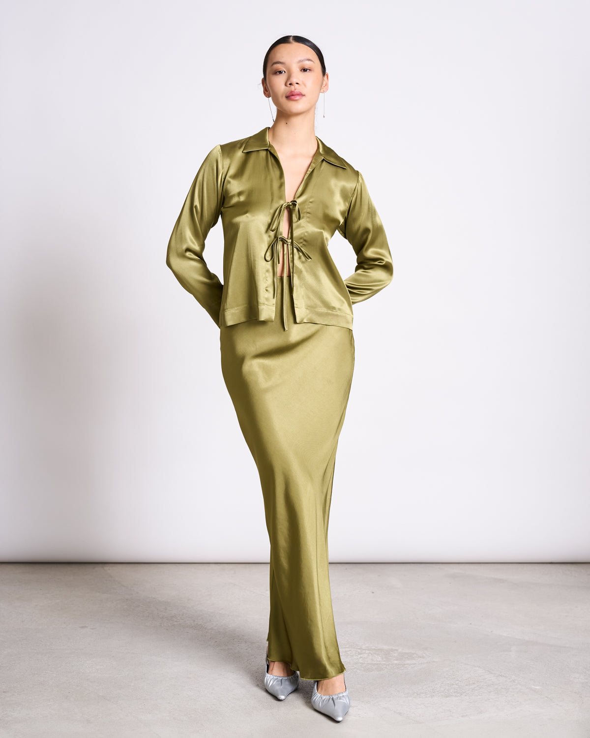 A woman poses against a plain backdrop wearing the jan-n-june TIE BLOUSE KALUZ WOODY OLIVE, styled with a matching long skirt made from ENKA® Viscose. Her hair is pulled back, and she wears pointed silver shoes to complete the look.
