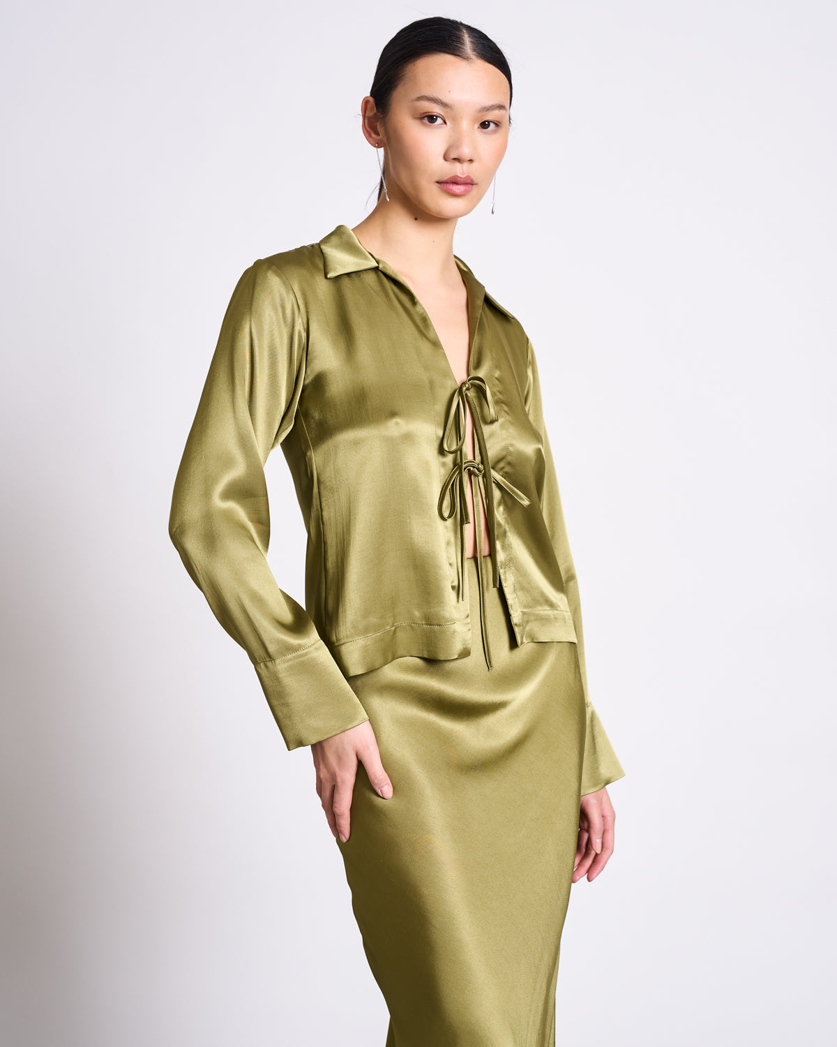 A woman wears the jan-n-june TIE BLOUSE KALUZ WOODY OLIVE, a shiny long-sleeve vegan silk blouse with a front tie, paired with a matching satin skirt, standing against a plain light background.