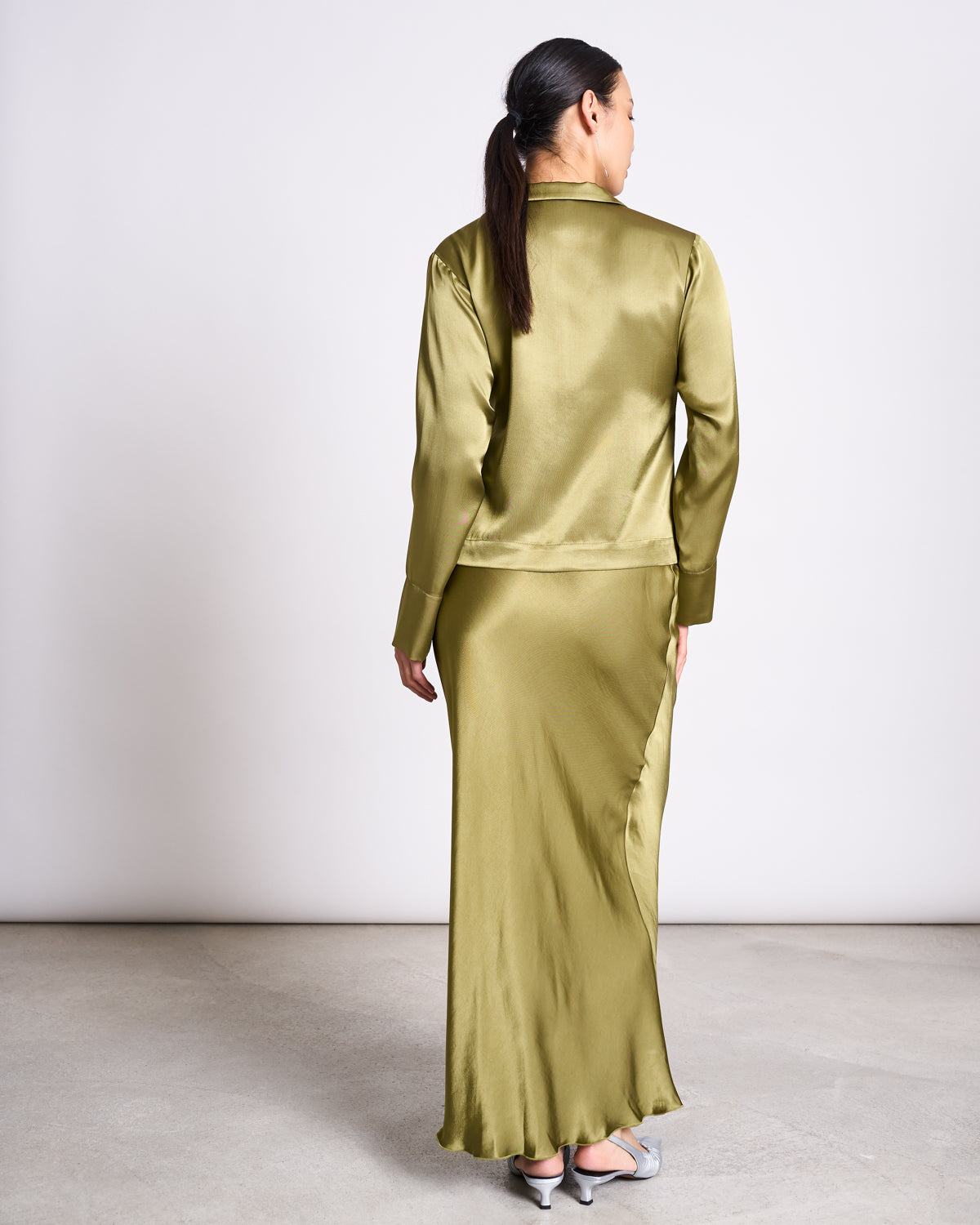 A woman with long dark hair in a ponytail stands facing away, wearing the jan-n-june TIE BLOUSE KALUZ in woody olive and a matching long skirt with light gray heels, against a plain light background.