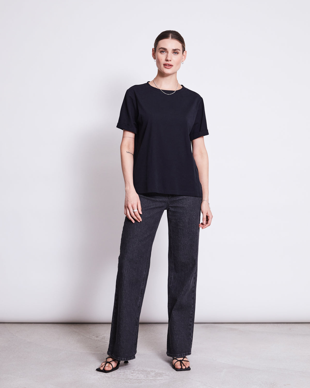 A woman stands against a white background wearing the jan-n-june BRUSHED T-SHIRT BOY BLACK GOTS, paired with loose fit wide-leg jeans and black heeled sandals. Her straight brown hair is pulled back as she looks at the camera neutrally.
