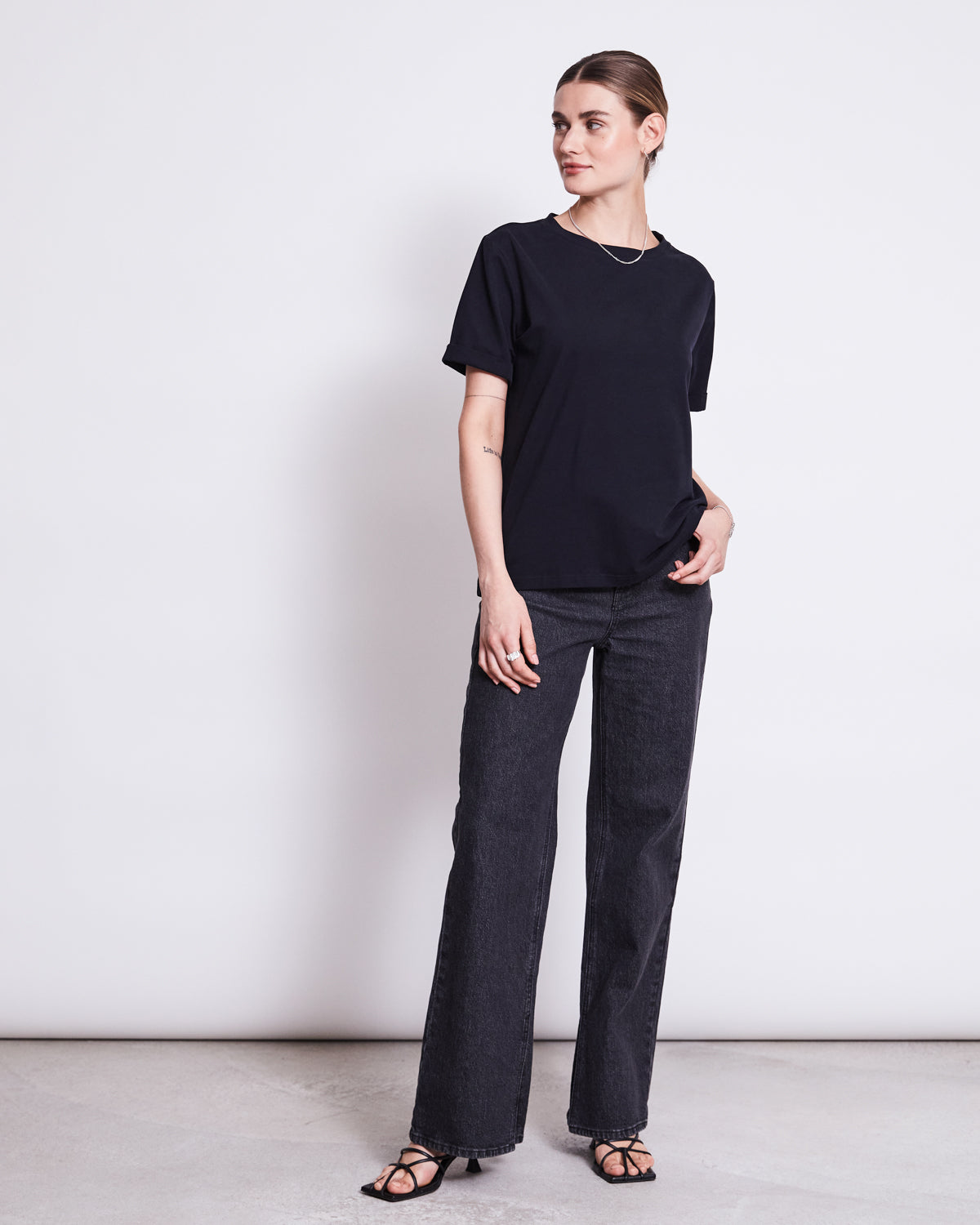 A woman stands against a white wall, looking to the side with her hair pulled back. She wears jan-n-junes BRUSHED T-SHIRT BOY BLACK GOTS, dark gray straight-leg jeans, and black heeled sandals.