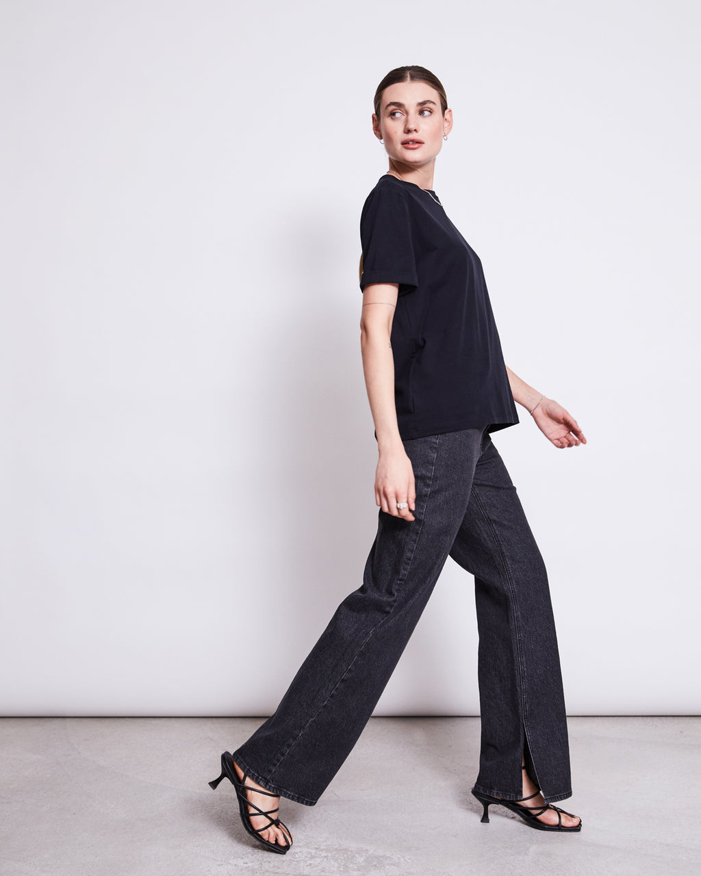 A woman in a jan-n-june BRUSHED T-SHIRT BOY BLACK GOTS, dark gray wide-leg jeans, and black heeled sandals walks sideways before a white wall, glancing over her shoulder with a neutral expression.