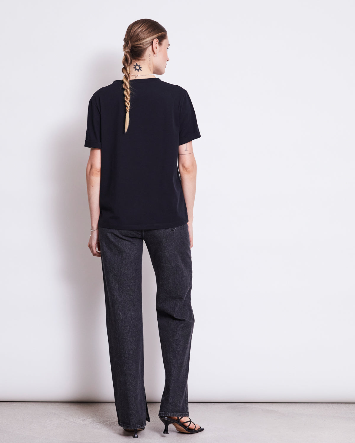 A person with a long braid and neck tattoo stands with their back to the camera, wearing the jan-n-june BRUSHED T-SHIRT BOY BLACK GOTS, dark jeans, and black heeled sandals against a plain white background.