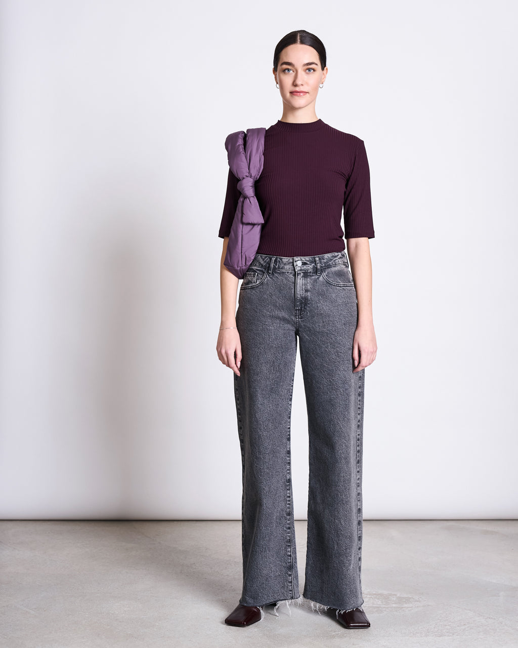 A woman stands against a plain light background in the jan-n-june LYOCELL RIB T-SHIRT NINA CHERRY LACQUER, gray high-waisted wide-leg jeans, dark shoes, with a purple jacket draped over her shoulder and her hair pulled back.