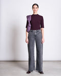 A woman stands against a plain light background in the jan-n-june LYOCELL RIB T-SHIRT NINA CHERRY LACQUER, gray high-waisted wide-leg jeans, dark shoes, with a purple jacket draped over her shoulder and her hair pulled back.