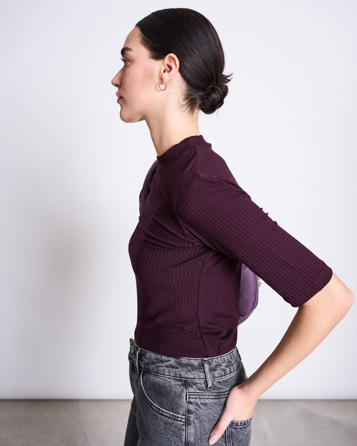 A woman with dark hair in a low bun stands in profile, wearing the jan-n-june TENCEL™ LYOCELL RIB T-SHIRT NINA CHERRY LACQUER and faded gray jeans against a plain light background.