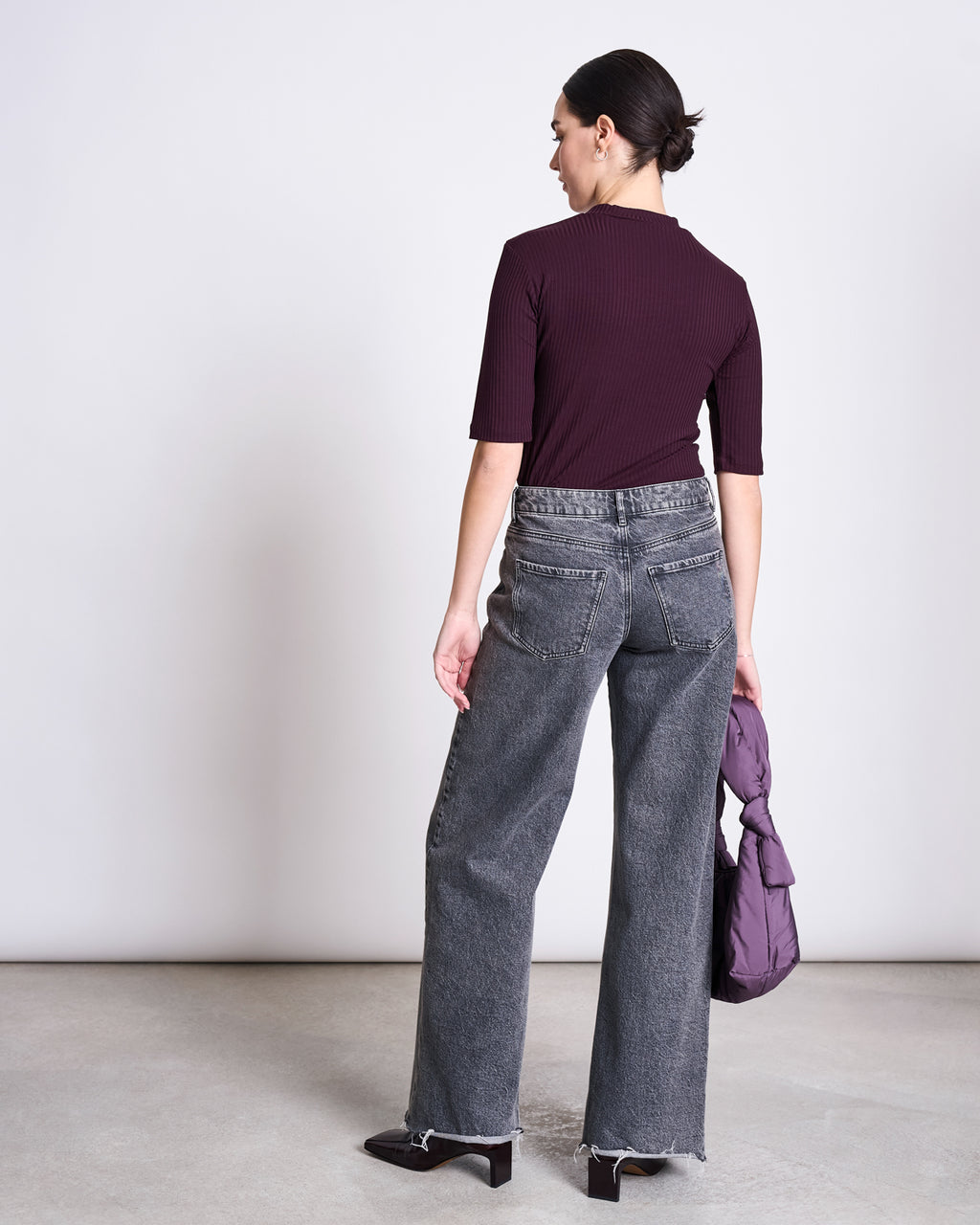 A woman faces away, wearing the jan-n-june TENCEL™ LYOCELL RIB T-SHIRT NINA CHERRY LACQUER, high-waisted gray jeans, and dark heels. She holds a purple puffer jacket in her left hand, her hair in a low bun.