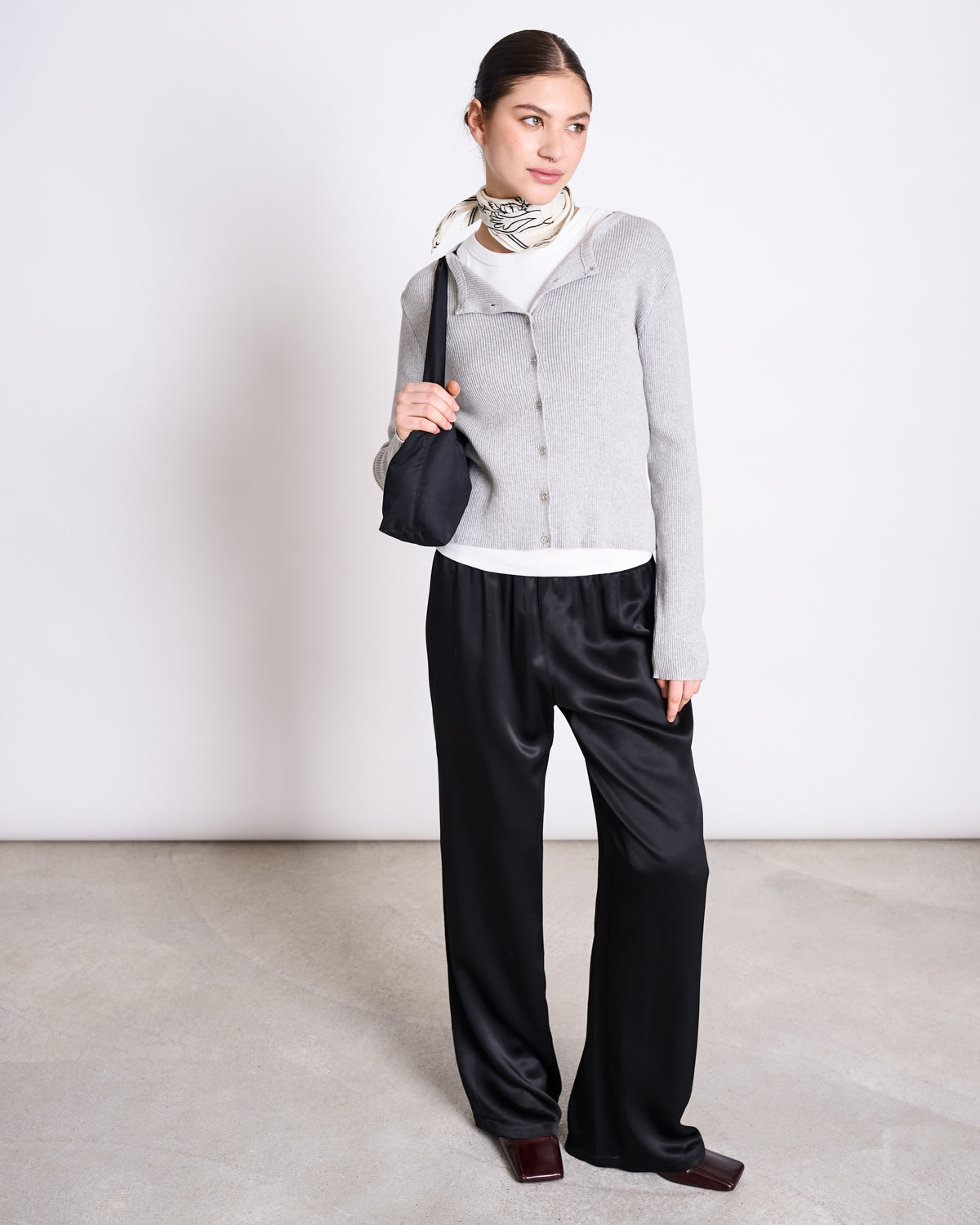 A woman stands against a plain background wearing the jan-n-june CREWNECK CARDIGAN GLIMT LIGHT GREY GOTS over a white top, black satin wide-leg pants, burgundy square-toed shoes, a patterned neck scarf, and holding a black shoulder bag.