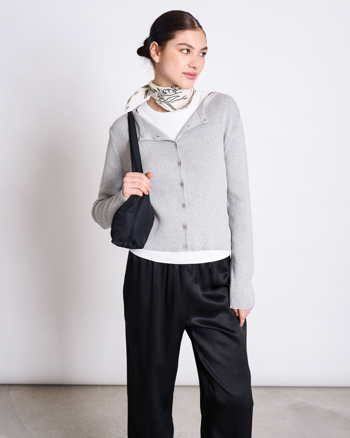 A woman stands against a plain background, wearing the jan-n-june CREWNECK CARDIGAN GLIMT LIGHT GREY GOTS over a white top, paired with black satin pants, a black shoulder bag, and a white scarf with a black pattern around her neck.
