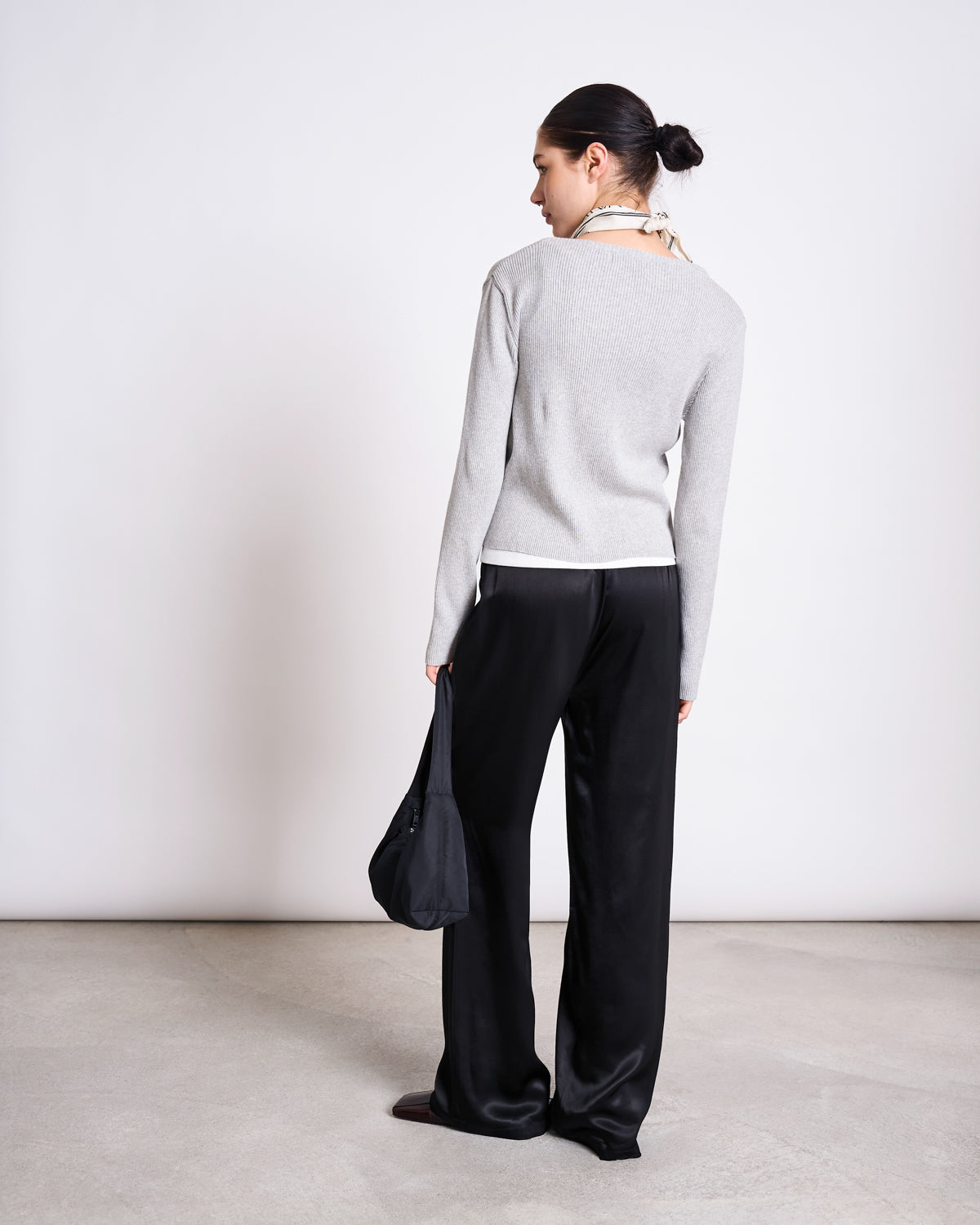 A woman with dark hair in a low bun faces away, wearing the jan-n-june CREWNECK CARDIGAN GLIMT LIGHT GREY GOTS, black wide-leg pants, a light scarf, and holding a black bag against a plain background.