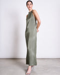 A woman stands sideways in the jan-n-june NECKHOLDER SLIPDRESS SICAYA SEA SPRAY—a sage green vegan silk maxi with a high neckline, open sides, and adjustable back ties—paired with silver strappy heels against a plain light background.