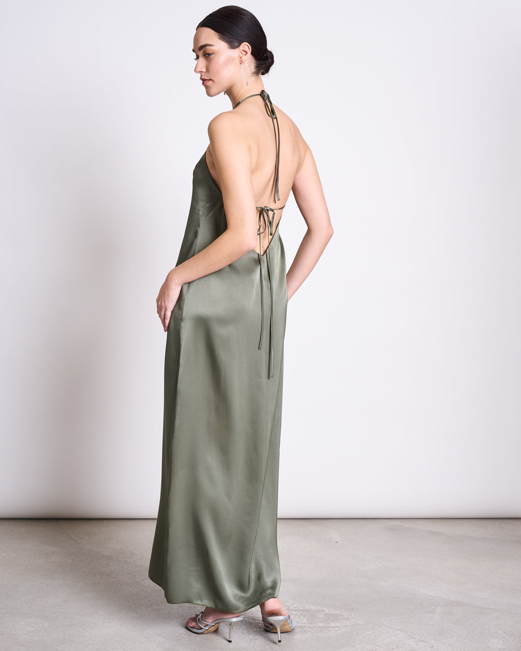 A woman in the jan-n-june NECKHOLDER SLIPDRESS SICAYA SEA SPRAY, a sage green halter-neck satin dress with open back and adjustable ties, stands on a light floor facing away, looking over her shoulder. Her hair is in a low bun and she wears strappy heels.