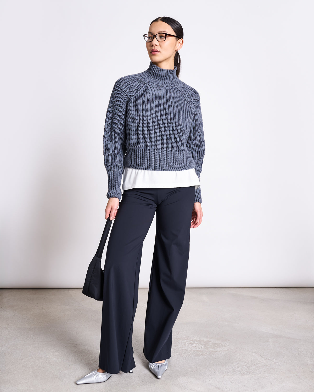 A woman in glasses wears the jan-n-june JUMPER OLA TURBULENCE GOTS over a white shirt with dark wide-leg trousers and silver pointed shoes, carrying a small black bag against a plain light background.