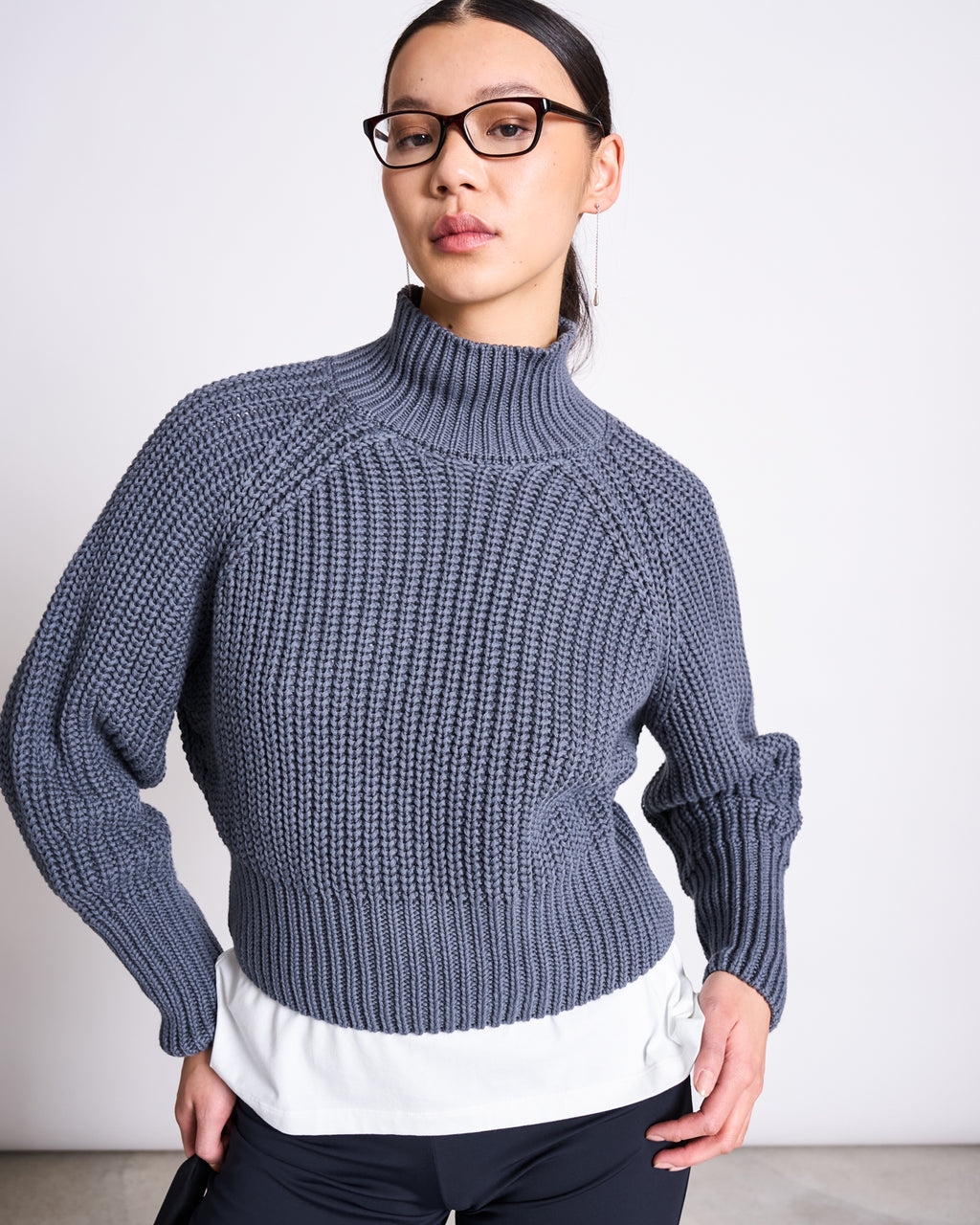 A woman in glasses wears the jan-n-june JUMPER OLA TURBULENCE GOTS—an oversized, chunky turtleneck jumper made from organic cotton—standing confidently with one hand on her hip against a plain background.