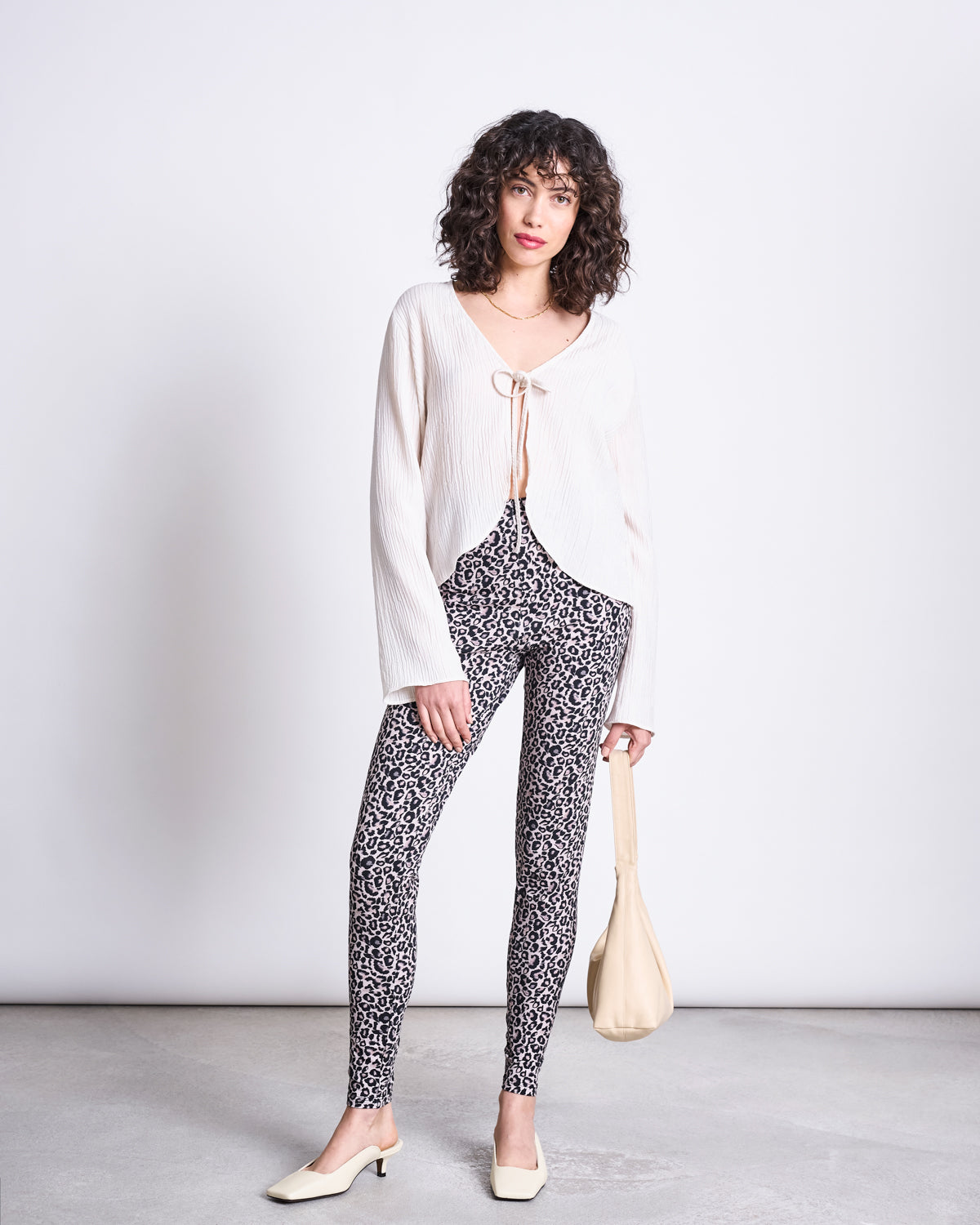A woman with curly hair wears jan-n-junes LEGGINGS LEO GOTS, a cream blouse, and holds a matching cream handbag. Her look is completed with cream low-heeled shoes, all set against a plain white background.