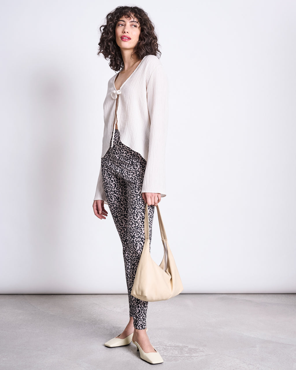 A woman with curly hair poses against a plain background, wearing jan-n-junes LEGGINGS LEO GOTS, a white textured cardigan, cream flats, and carrying a light beige shoulder bag.