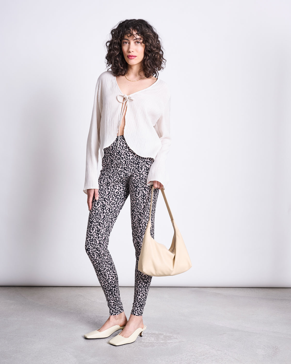 A woman with curly hair stands against a plain background, wearing a white long-sleeve blouse, jan-n-june LEGGINGS LEO GOTS, cream flats, and holding a beige shoulder bag.