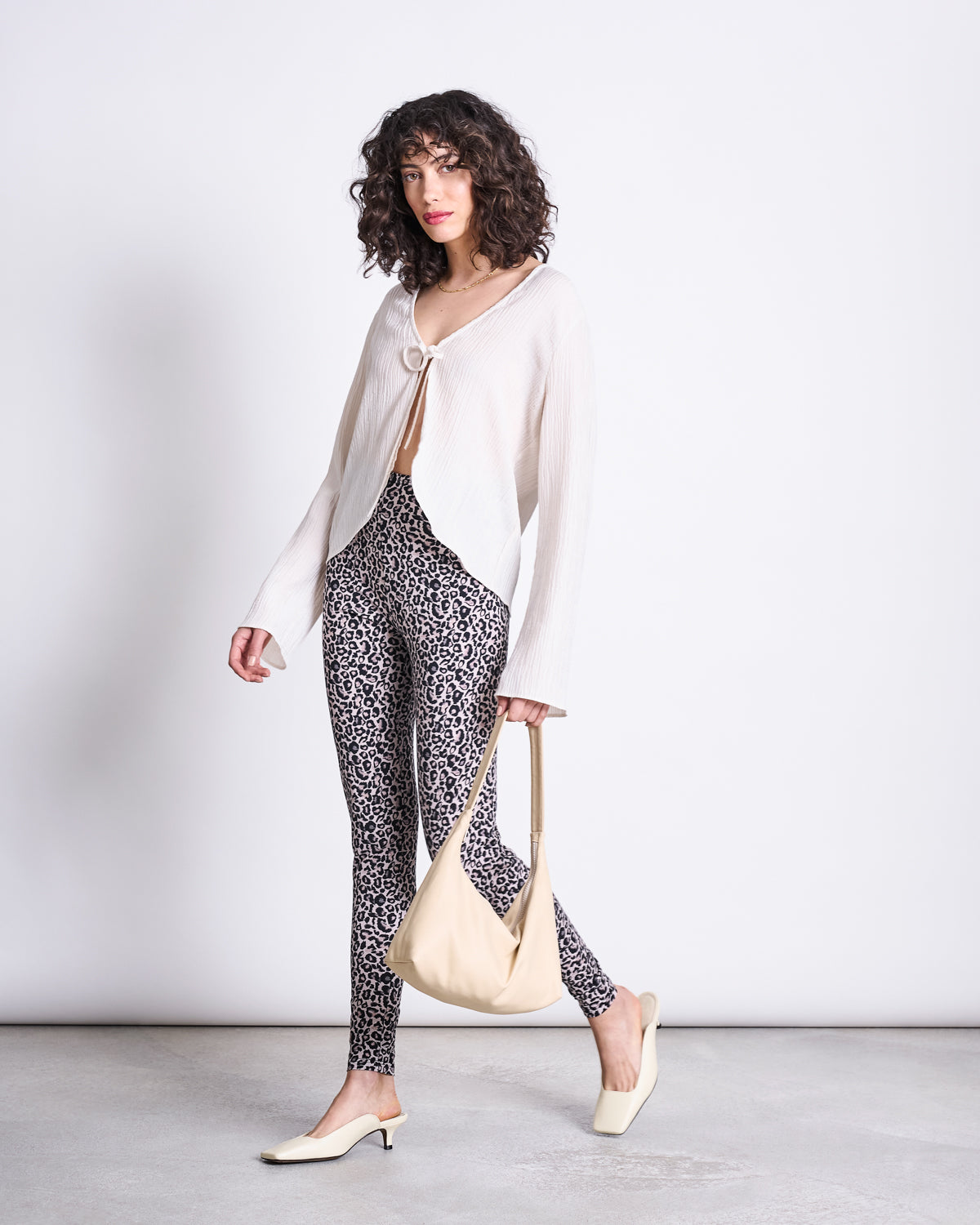 A woman with curly hair wears jan-n-junes LEGGINGS LEO GOTS, a high-waisted organic cotton leo print legging, paired with a white flowy long-sleeved top, cream heels, and a cream shoulder bag against a plain light background.