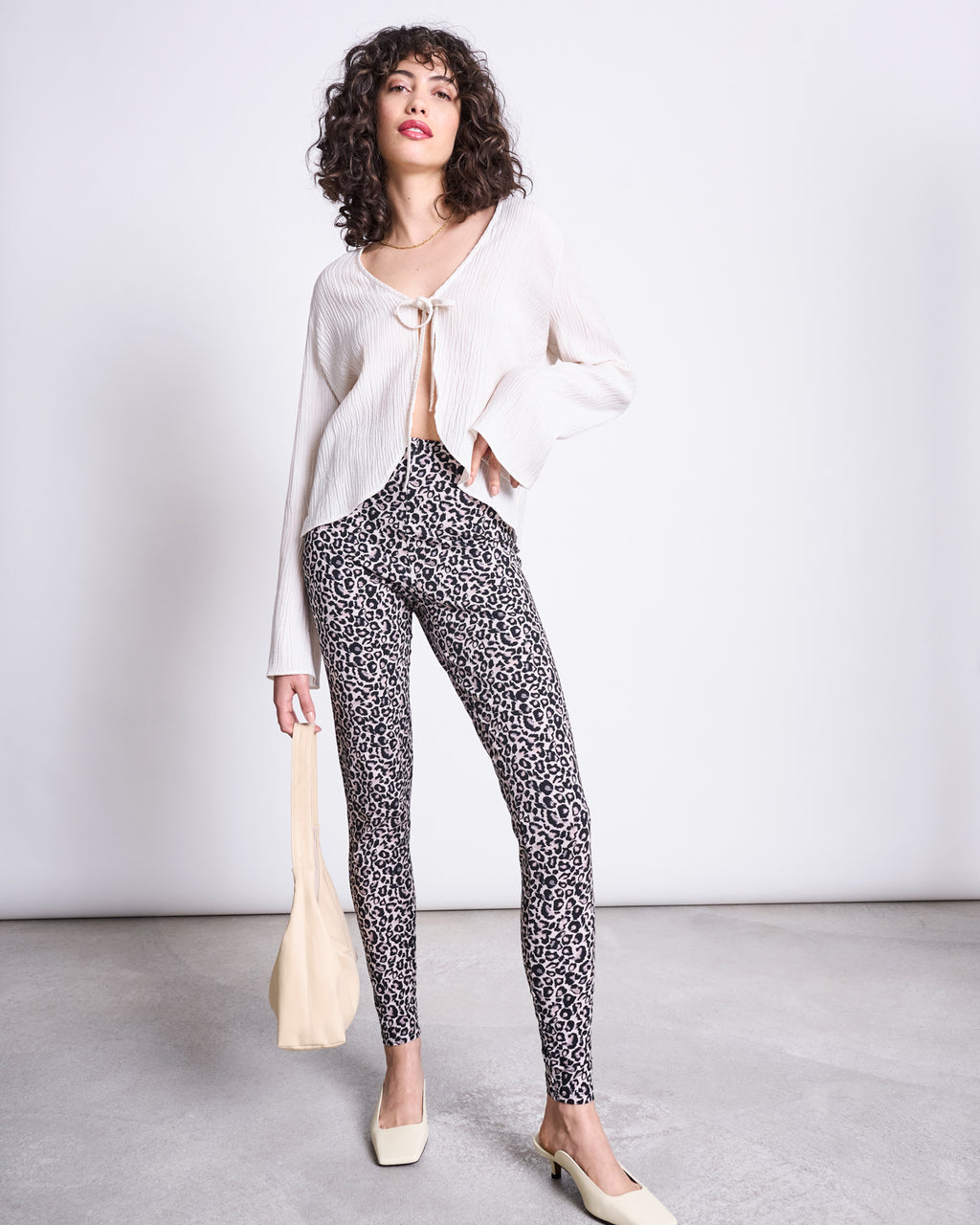 A woman poses confidently indoors, wearing jan-n-junes LEGGINGS LEO GOTS—high-waisted, organic cotton leo print leggings—paired with a cream blouse, square-toe shoes, and a beige bag against a plain background.