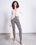 A woman poses confidently indoors, wearing jan-n-junes LEGGINGS LEO GOTS—high-waisted, organic cotton leo print leggings—paired with a cream blouse, square-toe shoes, and a beige bag against a plain background.