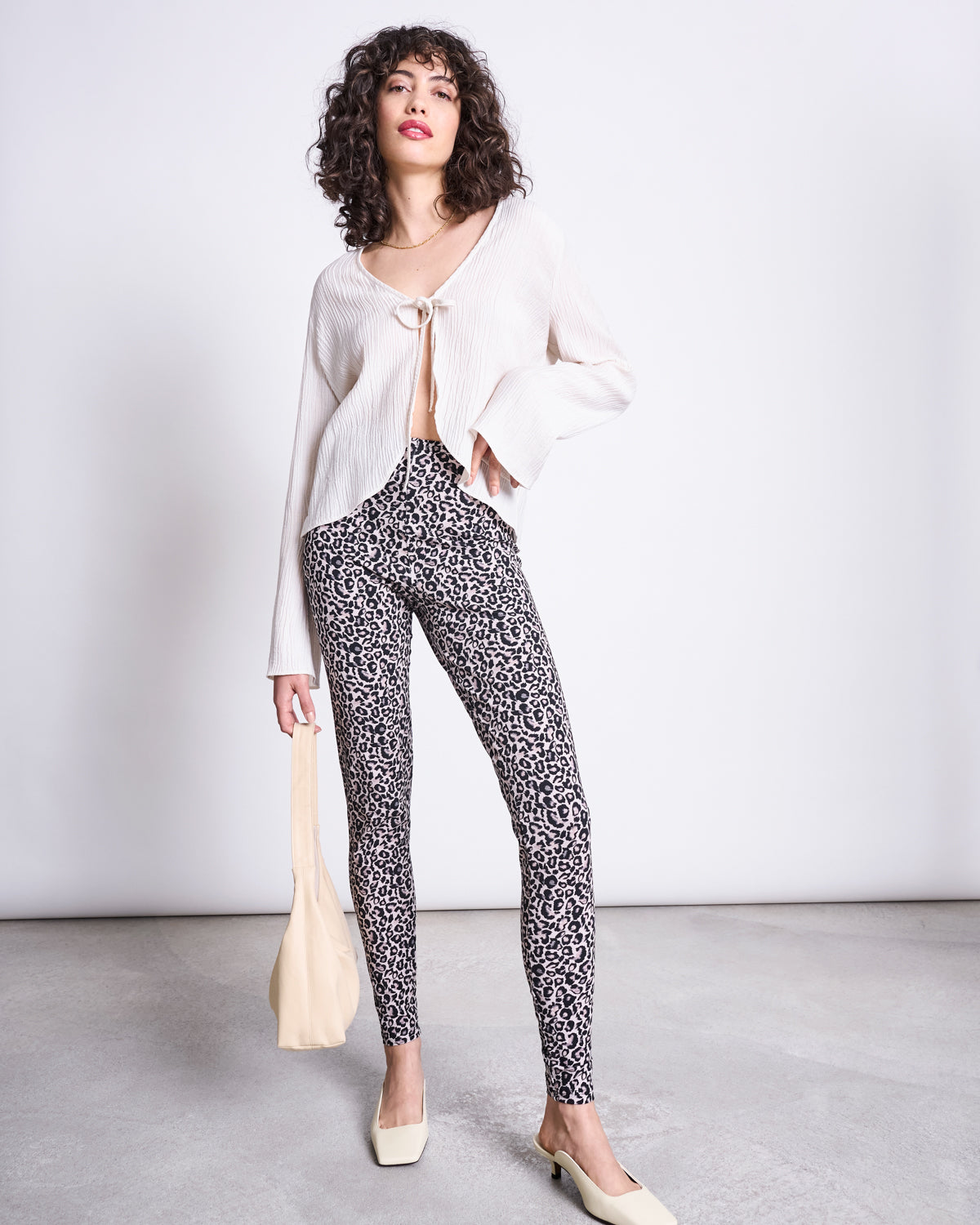 A woman poses confidently indoors, wearing jan-n-junes LEGGINGS LEO GOTS—high-waisted, organic cotton leo print leggings—paired with a cream blouse, square-toe shoes, and a beige bag against a plain background.
