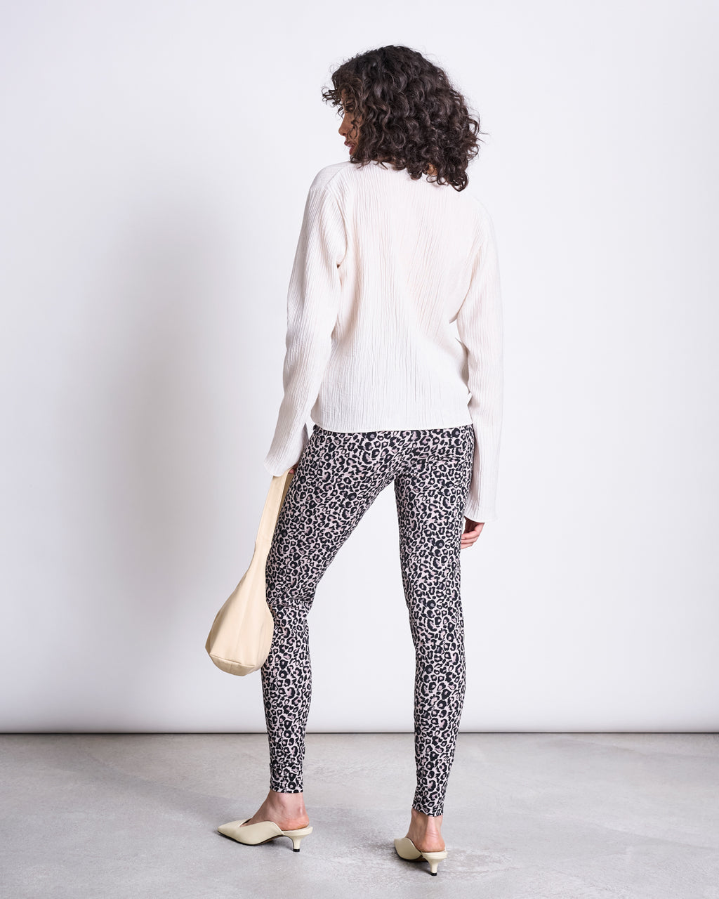 A woman with curly hair stands facing away, wearing a white ribbed sweater, jan-n-june’s LEGGINGS LEO GOTS in a high-waisted leo print, cream heeled shoes, and holding a light beige handbag against a plain light background.