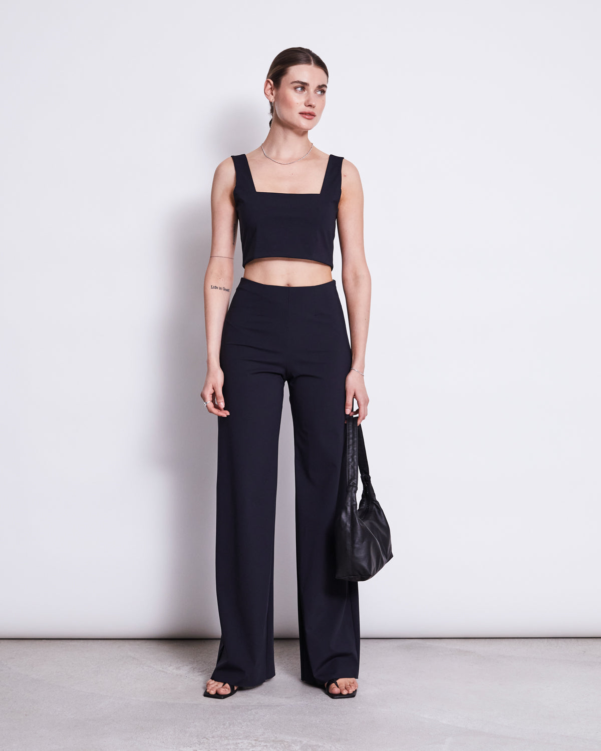 A woman stands against a white background wearing the jan-n-june CROP TOP CANCUN BLACK, a sleeveless square-neck crop top, paired with high-waisted black recycled polyamide trousers. She holds a black bag and her hair is pulled back.