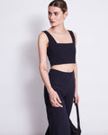 A woman models the jan-n-june CROP TOP CANCUN BLACK, a sleeveless crop top with a square neckline, paired with high-waisted black pants. She stands against a white background holding a black bag and looks left with a neutral expression.
