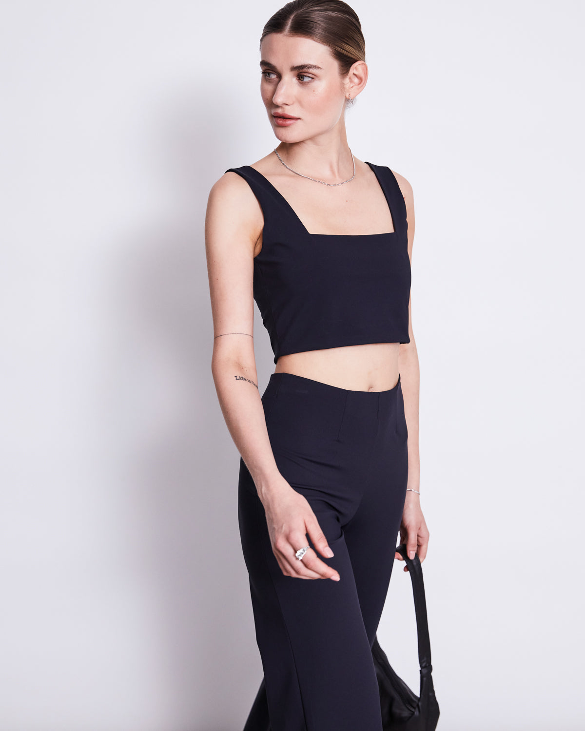 A woman models the jan-n-june CROP TOP CANCUN BLACK, a sleeveless crop top with a square neckline, paired with high-waisted black pants. She stands against a white background holding a black bag and looks left with a neutral expression.