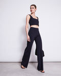 A woman with dark hair pulled back models jan-n-june’s WIDE PANTS MARLENE BLACK, made from sustainable fabric, styled with a black crop top and bag. She stands against a plain white wall, posing confidently as she looks to the side.