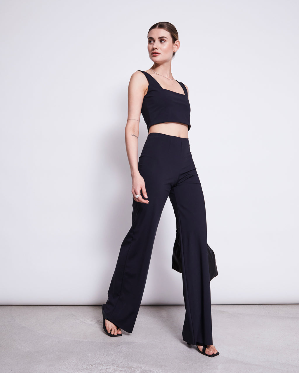 A woman poses against a plain white wall wearing the jan-n-june CROP TOP CANCUN BLACK, paired with high-waisted, wide-leg black pants, open-toe heels, and holding a black bag by her side.