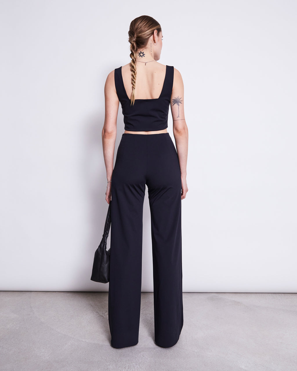 A woman with a braid, visible back tattoos, and a black handbag stands facing a white wall, wearing jan-n-june’s WIDE PANTS MARLENE BLACK high-waist pants and a black sleeveless crop top made from sustainable fabric.